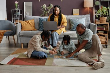 Dört kişilik aile, oturma odasında, yastık, bitki ve duvar süslemeleriyle dolu yerde yapboz faaliyetinde bulunuyor.