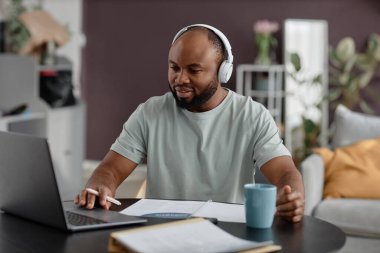 Masada oturan, kulaklık takan, dizüstü bilgisayarla ve belgelerle uğraşan, arka planda bitkiler ve rahat mobilyalarla rahatlayan bir adam.
