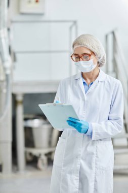 Maskeli ve önlüklü kadın işlem teknisyeninin ilaç imalat fabrikasındaki üretim kalitesini izlerken not defterine not aldığı dikey bir fotoğraf.