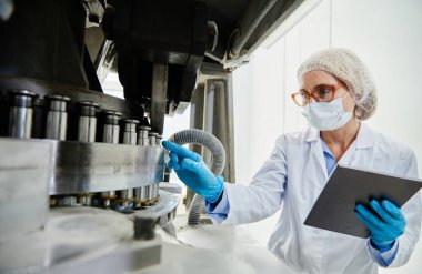 Profesyonel kadın işlem teknisyeni vinil eldivenler ve maske teftiş makineleri. İlaç imalat fabrikasındaki pres makinesinin yumruk aletlerine işaret ederken.