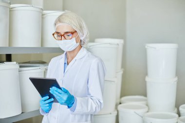 Konsantre kadın işlem teknisyeninin dijital tablet kullanarak büyük plastik kaplarla eczane deposunda envanter çıkarırken orta boy fotoğrafı.
