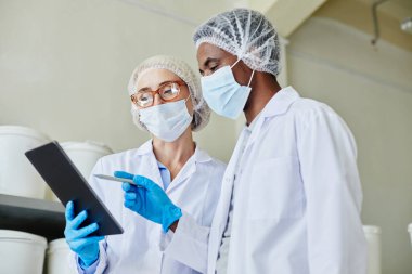 Kadın işlem teknisyeninin gözlük ve maske takıp Afro-Amerikan erkek iş arkadaşıyla dijital tablet kullanırken ilaç fabrikasındaki ürünlerin envanterini çıkarırken orta boy görüntüsü.