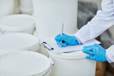 Mavi vinil eldivenli tanınmayan bir yapım işçisinin plastik konteynıra yazarken ilaç imalat fabrikasındaki not defterine not aldığı görüntüler.