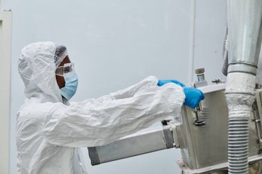Profesyonel Afro-Amerikan erkek teknisyen koruyucu tulumlar içinde fabrika ekipmanlarını korurken eczane zemininde üretim süreçlerini yürütüyor.