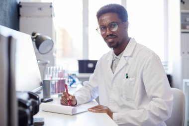 Laboratuvarda çalışma masasında oturmuş araştırma kayıtları hazırlarken kameraya gülümseyen Afrikalı Amerikalı erkek tıp bilimcisinin orta boy portresi.