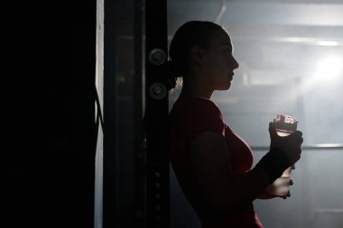 Spor salonunda hafif bir yıldırımla yapılan yoğun çalışmadan sonra elinde bir şişe spor içeceği tutan genç bir kadının silüeti.