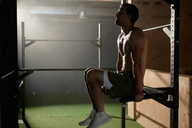 Paralel çubuklarda asılı kassız bir adam vücut kaslarını güçlendirmek için statik tutarak bacaklarını kaldırırken spor salonunda hafif bir yıldırımla çalışıyor.