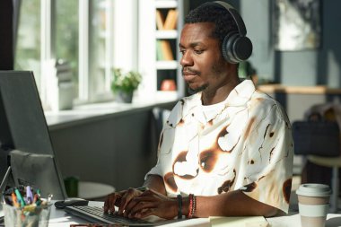 İş yerinde çalışan, teknoloji kullanan, fotokopi çeken ve büyük kulaklık takan yetişkin Afro-Amerikan bir adamın yan görüntüsü. 