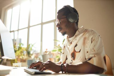 Genç siyahi bir adamın güneş ışığıyla aydınlatılan ofiste çalışırken bilgisayar kullanıp kulaklık taktığı sıcak ton portresi.