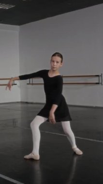 Geniş dans sınıfında kameraya zarif pozlarda poz veren siyah mayo ve beyaz tayt giyen genç balerinin dikey yavaş yavaş portresi.