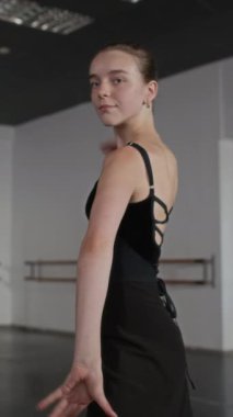 Siyah taytlı beyaz balerinin dikey portresi hazır duruş içinde modern stüdyoda kameraya gülümsüyor.