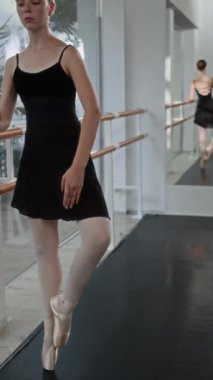 Dikey çekimde, siyah taytlı genç balerin, kadın eğitmenle barda dengeli hareketler yapıyor ve rehberlik ediyor.