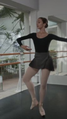 Siyah tayt ve beyaz taytlı beyaz balerin büyük pencereler ve bale çubuklarıyla stüdyoda dans gösterisi yapıyor.