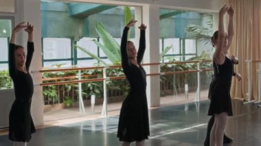 Bir grup genç beyaz bale dansçısı ve siyah tayt giymiş antrenörleri yerden tavana pencereli geniş güneşli stüdyoda prova yapıyorlar.