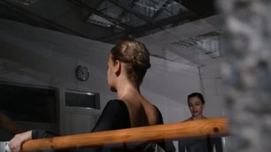 Geniş bale okulunda, bayan öğretmenler ders verirken, genç bale dansçıları ahşap barda esneme hareketleri uyguluyor.