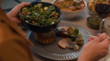 Tanınmayan bir kadın tabağa yeşil salata koyarken arkadaşlarıyla şenlikli masada kırmızı şarap ve aromalı yemeklerle şenlikli bir yemek yiyor.