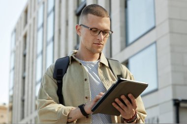 Gözlüklü bir adamın orta boy portresi iş yerine giderken veya dışarıda çalışırken tablet bilgisayar kullanarak ekrana bakması, uzayı kopyalaması