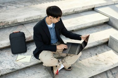 Kulaklıklı erkek bilgisayar geliştiricisinin şehir sokağındaki beton basamaklarda çalışırken çektiği yüksek açılı görüntüler. Boşluğu kopyala