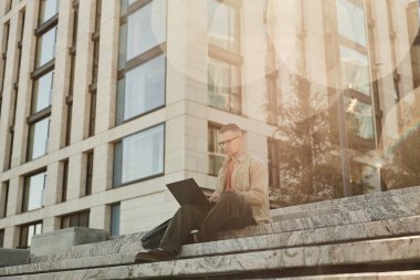 Şehir caddesindeki iş binasının önündeki beton basamaklarda otururken dizüstü bilgisayarı kullanarak kod yazmaya çalışan erkek bilişim programcısının tam karesi, bokeh etkisi, boşluk kopyalama