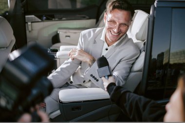 Süper starların özel yorumlarını alan gazeteciler havalı bir tonda çekilirken, televizyon medyasıyla konuşurken, arabanın arka koltuğunda gülümseyen erkek ünlünün orta boy görüntüsü.