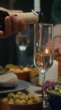 Akşamki Noel kutlaması için akşam yemeği masasında, ekose gömlekli, bardağa köpüklü şarap dolduran, tanınmayan birinin dikey yakın çekimi.