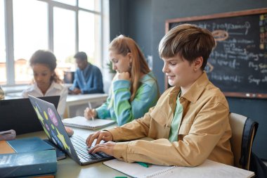 Modern okul sınıfının fotokopi alanının keyfini çıkarırken dizüstü bilgisayar kullanan gülümseyen genç çocuğun yan görünümü