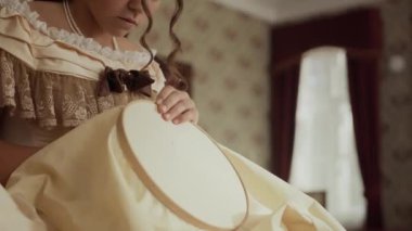 Klasik elbise ve inci kolye giyen genç aristokrat bir kadının 19. yüzyılın zarif salonlarında çiçek duvarları ve süs perdeleri olan dikiş işi yaparken çekilen pozu.