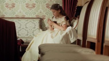 19. yüzyılın zarif, klasik elbiseli genç hanımı, gün içinde dikiş dikerken ya da nakış işlerken çiçekli duvarlar ve iplik iğneleriyle döşenmiş koltukta oturuyor.