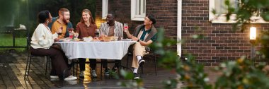 Wide angle shot of diverse group of adult people enjoying dinner party together at outdoor patio by brick house copy space clipart