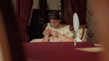 19. yüzyıl giysileri giymiş, kırmızı perdeli, masa örtülü ve klasik dekorlu eski moda bir odada dikiş dikmeye çalışan konsantre bir kız.