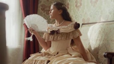 19. yüzyıldan kalma güzel elbiseli genç bir hanımın, güneşli oturma odasındaki pahalı döşeme kanepesinde elinde yelpazeyle otururken hayal kurduğu orta boy bir fotoğraf.