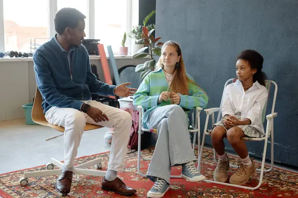 Yetişkin Afro-Amerikan bir adamın erkek öğretmen ya da akıl hocası olarak iki genç kıza stüdyoda yardım eden ve yaratıcı projeyi yönlendiren tam boy portresi.