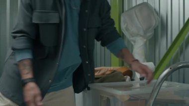 Kafkasyalı bir adam modern metal duvarlı depolama ünitesinde karton kutular, plastik konteyner ve bavulu el arabasına yüklüyor.