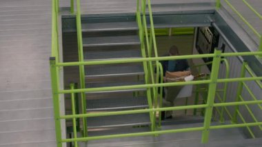 Kafkasyalı bir adamın elinde kutuyla modern depolama tesisinde canlı yeşil parmaklıklarla merdivenleri çıkarken çekilen fotoğrafı.