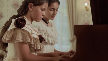 İki zarif 19. yüzyıl kadınının klasik dekorlu ve çiçekli güzel döşenmiş odada piyano dersi verirken orta açıdan çekilmiş bir fotoğrafı.