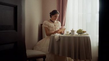 Klasik dantel elbiseli romantik bir kadın Viktorya dönemi dekorunda pencere kenarında oturuyor, el yazısıyla aşk mektubu okuyor ve gülümsüyor.