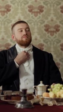 Beyaz kravatı düzeltirken kameraya bakan genç sakallı aristokratın dik bel portresi. Gündüz vakti klasik dekore edilmiş yemek odasında oturuyor.