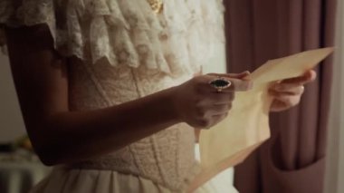 Klasik Viktorya dönemi elbisesi giymiş, kağıt üzerinde el yazısıyla mektup okuyan ve odasının penceresinde dikilirken göğsüne yakın duran genç bir kadının fotoğrafını çek.