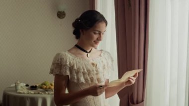 Orta boy beyaz kadın, dantelli Viktorya dönemi elbisesi giymiş, elinde aşk mektubuyla pencerede duruyor, okuyor, göğsüne yakın tutuyor ve şefkatle gülümsüyor.