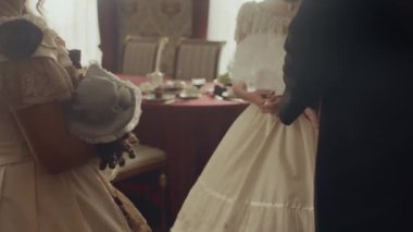 Viktorya döneminden kalma bir beyefendi ve iki kadın şık takım elbiseli resmi yemek masasının yanında durmuş, elinde şapka ve yelpaze tutarak sohbet ediyorlar.