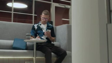 Odaklanmış genç Kafkasyalı serbest çalışan ya da gözlüklü öğrenci cep telefonlarını kontrol ederken, modern çalışma alanlarında masada otururken,