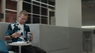 Gözlüklü, siyah kollu, kısa saçlı, akıllı telefon kullanan, kahvesini yudumlayan ve modern çalışma alanında çalışırken not defterine not alan beyaz bir adamın orta boy fotoğrafı.