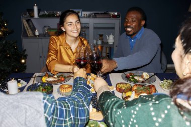 Medium shot of cheerful woman and Black man clinking wine glasses celebrating Christmas with friends during festive dinner at home party, camera flash clipart