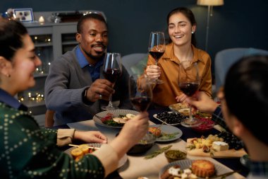 Orta ölçekli, neşeli, elinde şarap kadehiyle kadeh kaldıran siyahi adam ve kadının Noel 'i arkadaşlarıyla birlikte evde kutlama masasında otururken çekildiği bir sahne.