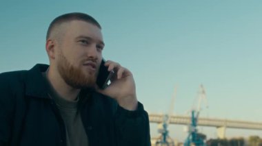 Kafkasyalı sakallı dedektifin endüstriyel arka planda cep telefonuyla konuşurken çekilen fotoğrafı ceketinin altında gizli bir el ile.