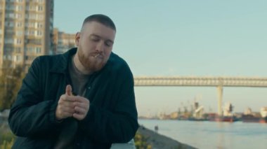 Beyaz sakallı polis memuru ya da sivil kıyafetli dedektif sigara içip ipuçlarını düşünürken arka planda gündüz manzarası olan rıhtım kenarındaki korkuluklara yaslan.