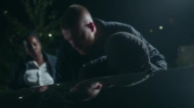 Afrika kökenli Amerikalı bayan meslektaşı olan profesyonel bir polis memuru maskeli ve kelepçeli şüpheliyi gece gözetimi sırasında araba kaputuna kafa üstü yatarken arıyor.