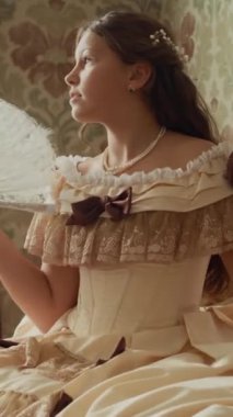 19. yüzyıldan kalma güzel elbiseli genç bir hanımın dikey çekimi. Güneş ışığıyla döşenmiş oturma odasındaki pahalı döşemelerde yelpaze ile hayal kurarken.