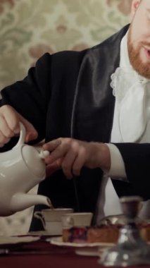 19. yüzyıldan kalma zarif bir beyefendinin porselen bardağa çay dökerken çay masasında oturduğu dikey belden yukarı pozu.