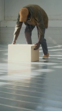 Genç siyahi adamın dikey portresi karton kutuyu yerden kaldırıyor, güneş ışığıyla aydınlanan geniş bir depoda duruyor ve kameraya bakıyor.